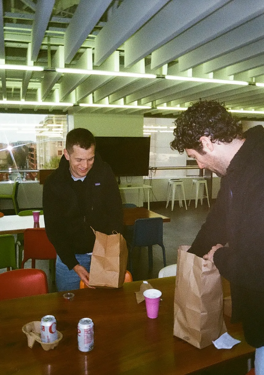 Two teammates unpacking takeout bags onto a table in the office kitchen.
